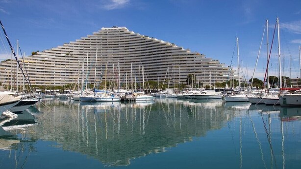 A marina with multiple boats docked in front of a large, modern hotel with a unique, tiered design. - Cocon Marina—Baie des Anges (Villeneuve-Loubet)