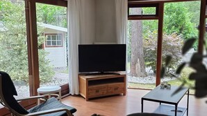 A room with a television on a wooden stand, a dining table with plates and glasses, and large windows offering a view of greenery and a house.