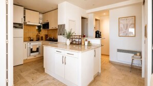 A modern kitchen with white cabinetry, a central island, and a dining area with a radiator. - 3-Bedroom Apartment with Panoramic Ocean View · 100 m² Terrace · Marina (Villeneuve-Loubet)