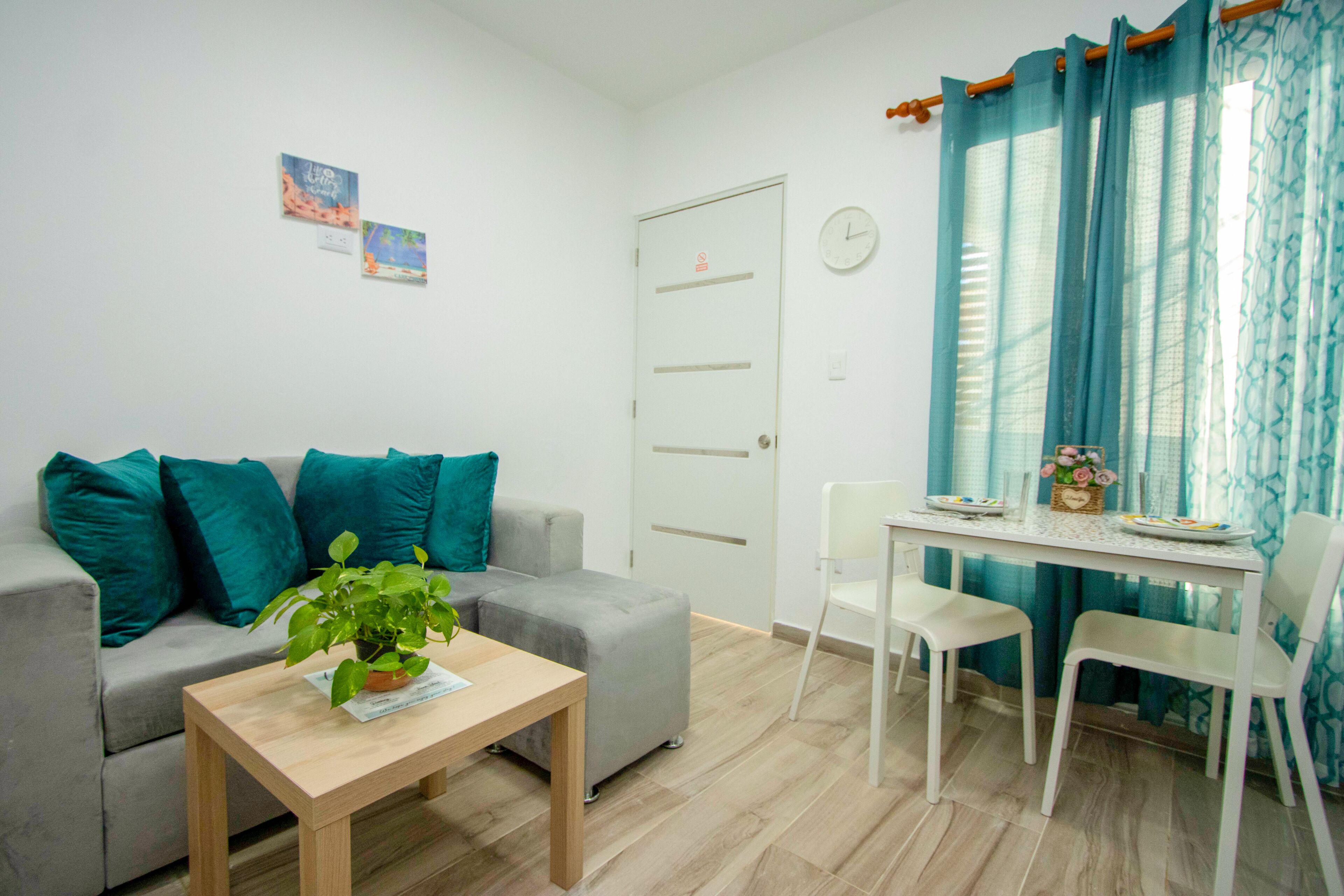 Una sala de estar con un sofá, una mesa pequeña con una planta, una mesa de comedor con sillas y una puerta.