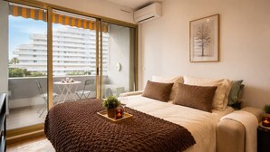 A bedroom with a bed, a bedside table, a window with a view of a building, and a balcony with chairs. - Ocean and Marina View · Terrace · Sleeps 4 · Riviera (Villeneuve-Loubet)