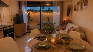 A dining area with a round table set for four, a sofa, a TV, and a view of the outdoors. - 2-Bedroom Apartment with Private Terrace · Pool · 50m from the Beach · Parking (Villeneuve-Loubet)