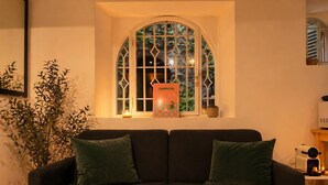 Un sofá oscuro con dos cojines verdes, una ventana con un marco decorativo y una mesita con una vela y una caja etiquetada 'COSMOPOLITAN'.