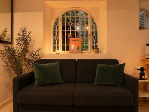 Ein dunkelgraues Sofa mit zwei grünen Kissen, ein Fenster mit dekorativem Rahmen und ein kleiner Tisch mit einer Kerze und einer Schachtel mit dem Aufdruck 'COSMOPOLITAN'.