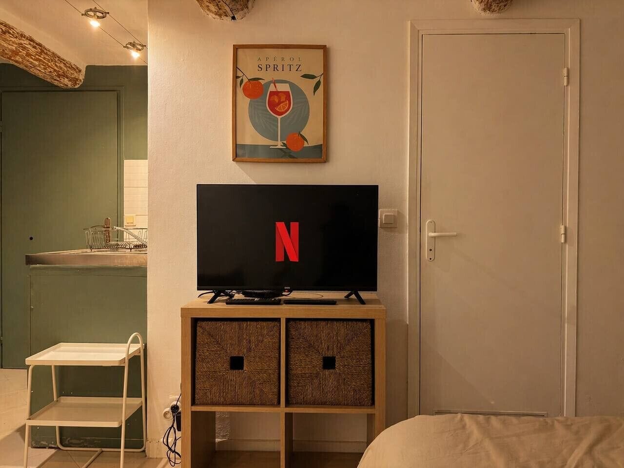 Une pièce avec un téléviseur affichant le logo de Netflix, une affiche encadrée au-dessus du téléviseur, une porte et une petite étagère blanche.
