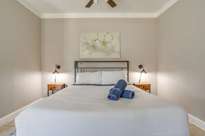 A neatly made bed with white linens and blue towels, two bedside tables with lamps, a ceiling fan, and a floral artwork above the bed. - Spacious Dallas Home + Guest House | Sleeps 21 (Dallas)
