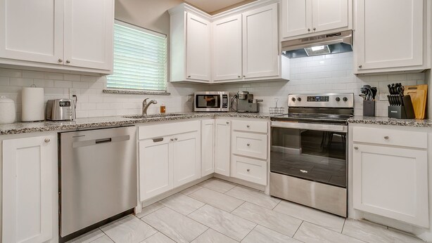 A modern kitchen with white cabinets, a dishwasher, a microwave, a stove, and a window with blinds. - Spacious Dallas Home + Guest House | Sleeps 21 (Dallas)