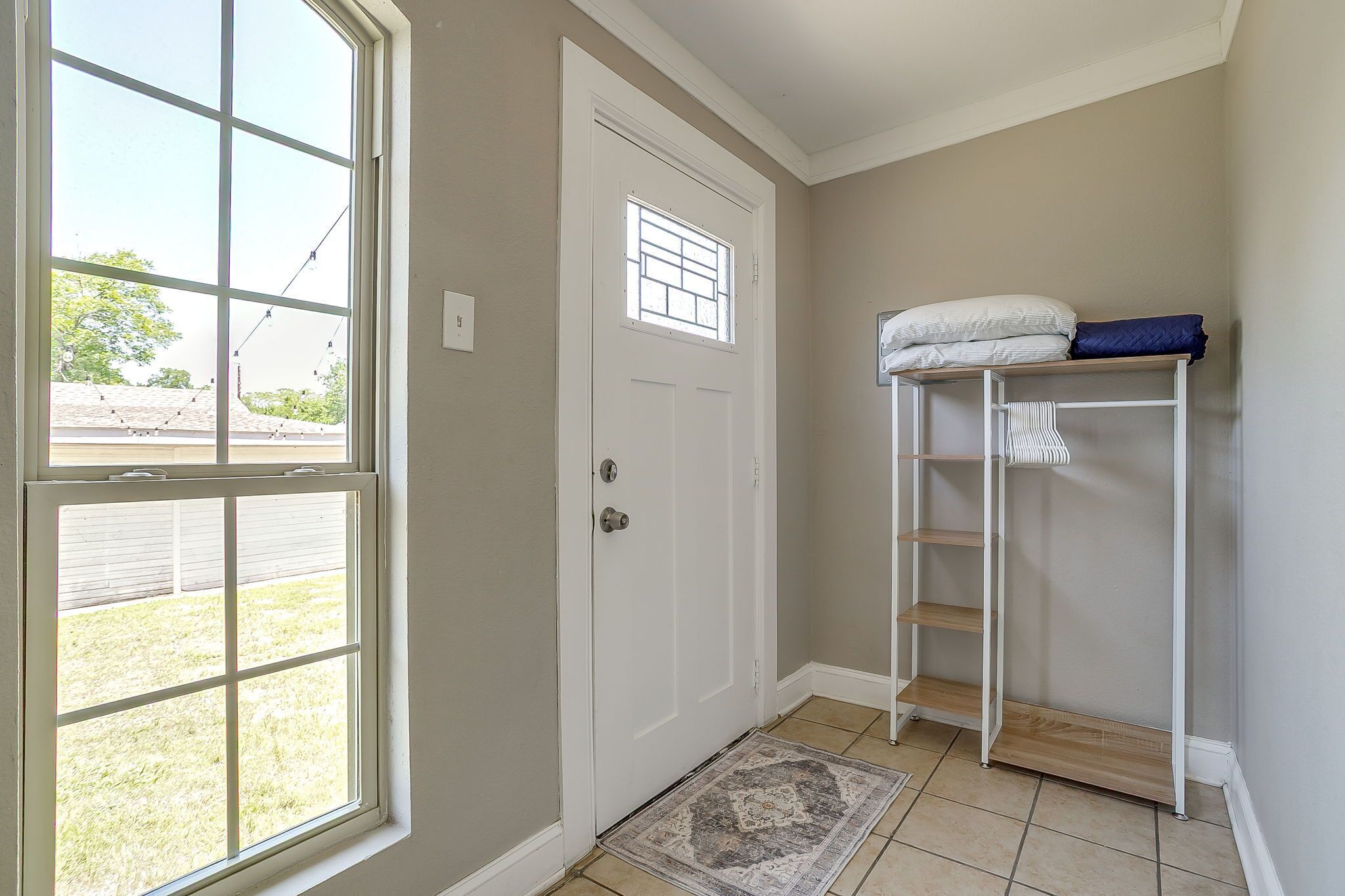 A room with a white door, a window, a small rug, and a white metal shelf with a folded blanket.