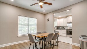 A dining area with a table and chairs, a ceiling fan, a window with blinds, and a view into a kitchen area. - Spacious Dallas Home + Guest House | Sleeps 21 (Dallas)