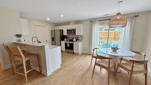 A modern kitchen with white cabinets, a dining area with a round table and chairs, and a large window with a view of the outdoors. - Fully Furnished Brand-New 3BR/2.5BA Home ~ Hot Tub ~ Close to Airport (Wilmington)