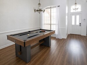 A pool table with balls, a chandelier, and a door in a room with wooden flooring and white walls. - Pool + Hot Tub | DFW Airport + Stadiums (Euless)