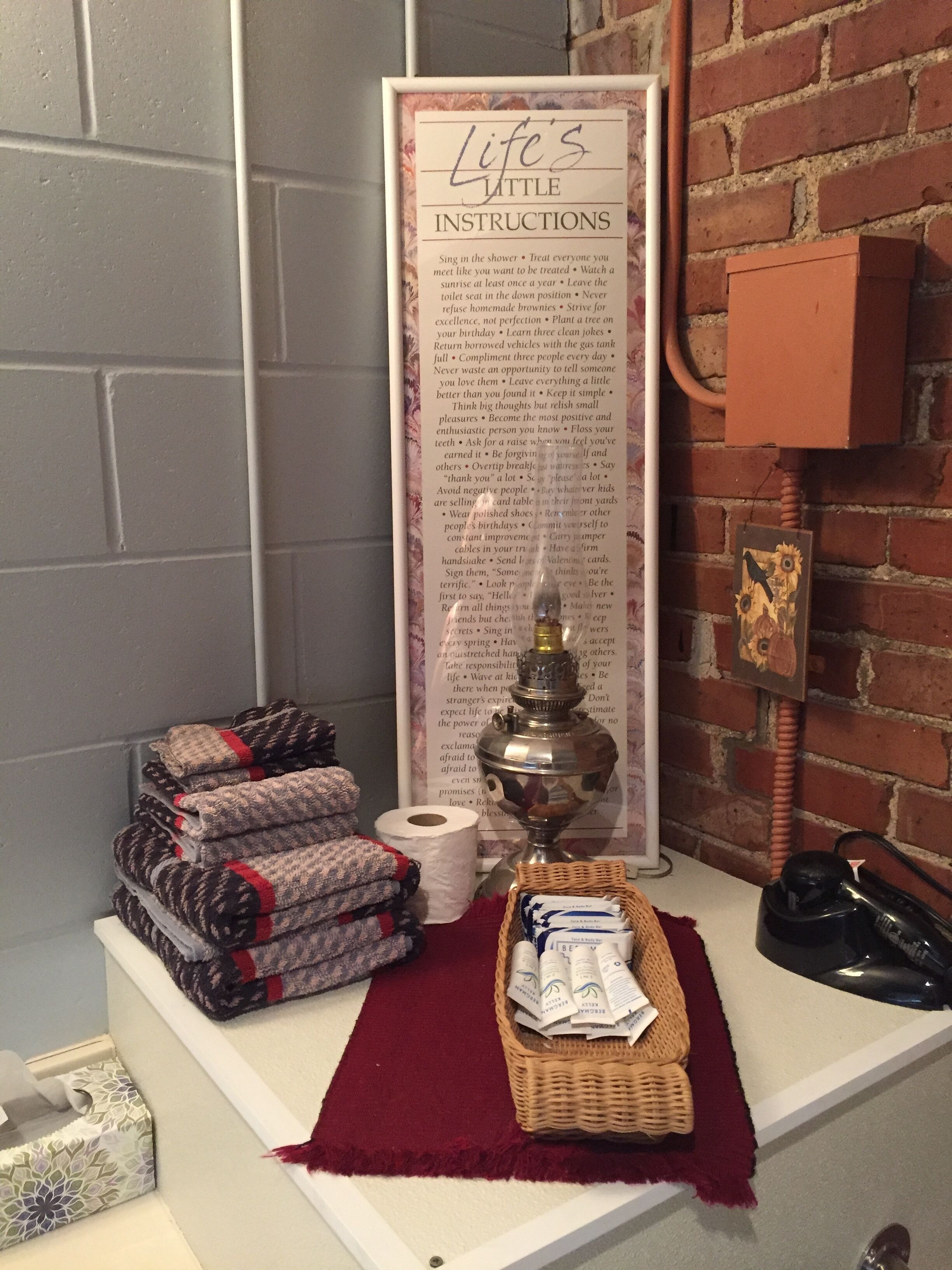 A bathroom with a sign listing 'Life's Little Instructions', towels, a roll of toilet paper, a lamp, and a telephone.