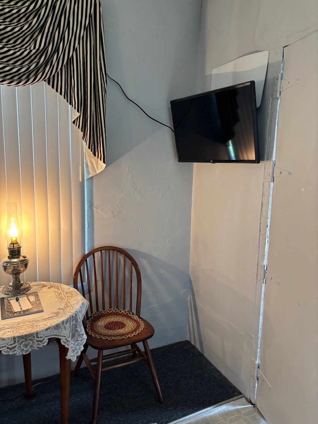 A room with a television mounted on the wall, a wooden chair, a table with a lace tablecloth, and a lamp.