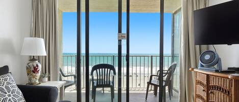 A room with a sliding glass door leading to a balcony with beach views, a television, a lamp, and a wicker cabinet.
