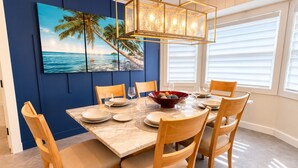 Un coin repas avec une table dressée pour quatre personnes, des chaises, un bol de collations et un tableau représentant une scène de plage accroché au mur.