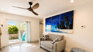 Un salon comprenant un canapé, un ventilateur de plafond, un tableau représentant une scène de plage et une porte-fenêtre coulissante donnant sur une terrasse avec des chaises.
