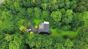 An aerial view of a house surrounded by dense green forest, with a driveway leading to the entrance.