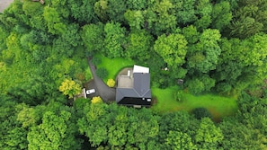 An aerial view of a house surrounded by dense green forest, with a driveway leading to the entrance.