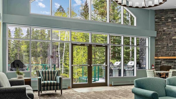 A spacious living room with large windows, a stone fireplace, and a view of the outdoors. - Cozy 1-bedroom condo in gorgeous Canmore with Indoor/Outdoor Pool and Spa (Canmore)