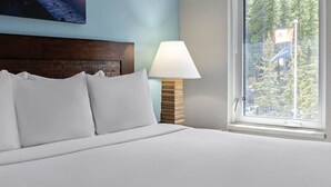 A hotel room with a bed, a wooden headboard, a lamp, and a window with a view of trees and a flag. - Cozy 1-bedroom condo in gorgeous Canmore with Indoor/Outdoor Pool and Spa (Canmore)