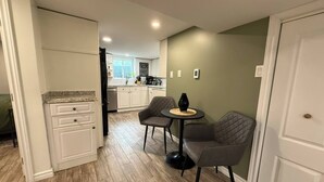 A compact kitchen with white cabinets, a dining area with two chairs and a small table, and a door leading to another room.