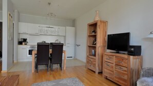 A modern kitchen with white cabinets, a dining area with a table and chairs, a wooden entertainment unit with a TV, and a gray rug on the floor. - Exclusive 2-room apartment in a prime location on Neumarkt (Dresden)
