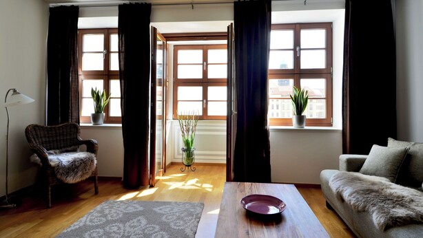A room with a sofa, a chair, a table, a lamp, and two windows with curtains. - Exclusive 2-room apartment in a prime location on Neumarkt (Dresden)