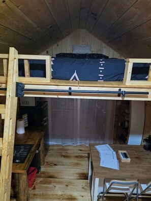 A wooden bunk bed with a desk and chair underneath, a small kitchen area, and a window with curtains.
