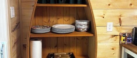 A wooden boat-shaped shelf unit with multiple compartments, including a section for plates and bowls, a section with black containers, and a section with a roll of paper towels.