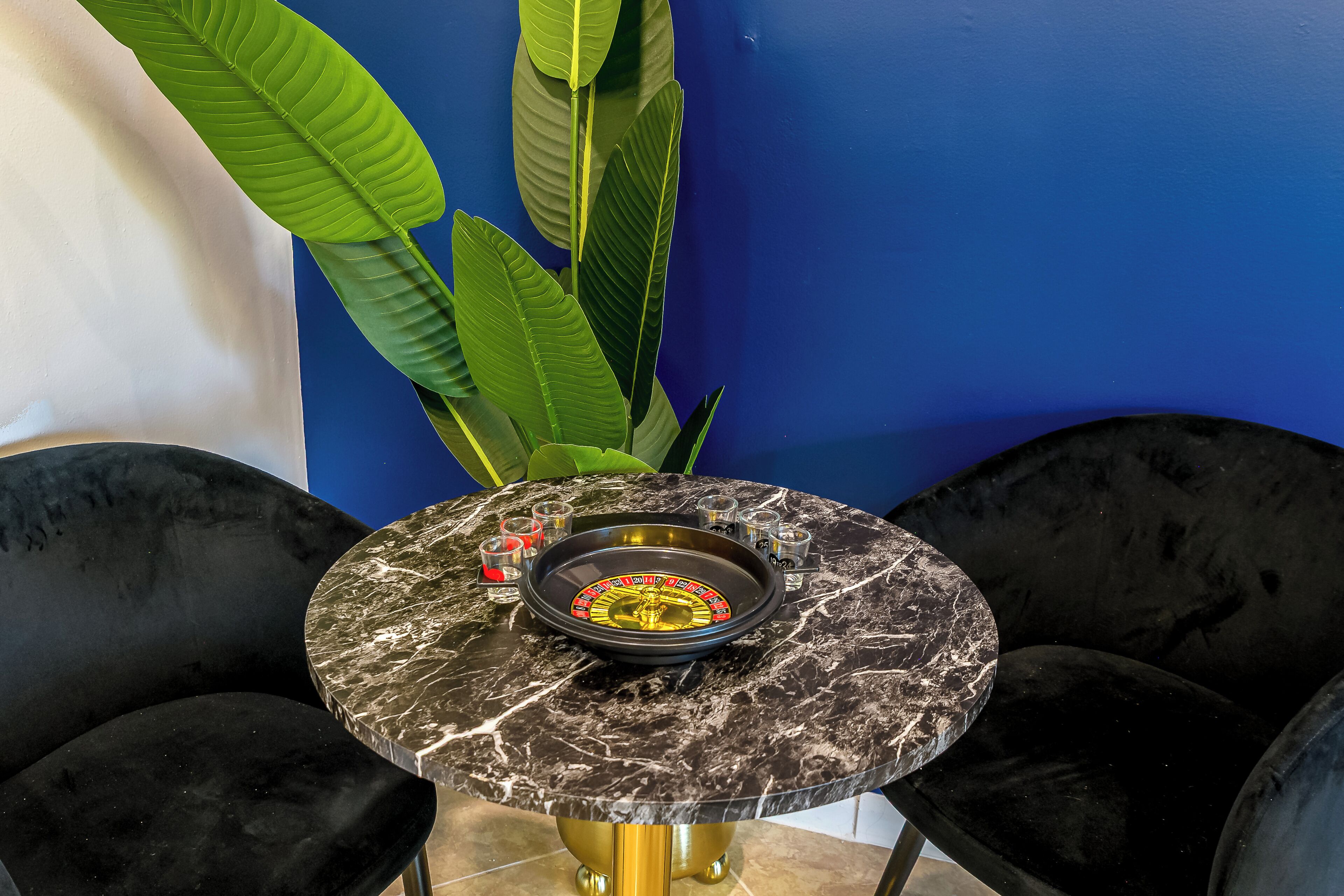 Une table ronde surmontée d’un plateau en marbre, entourée de chaises noires, le tout placé devant un mur bleu orné de grandes plantes vertes.