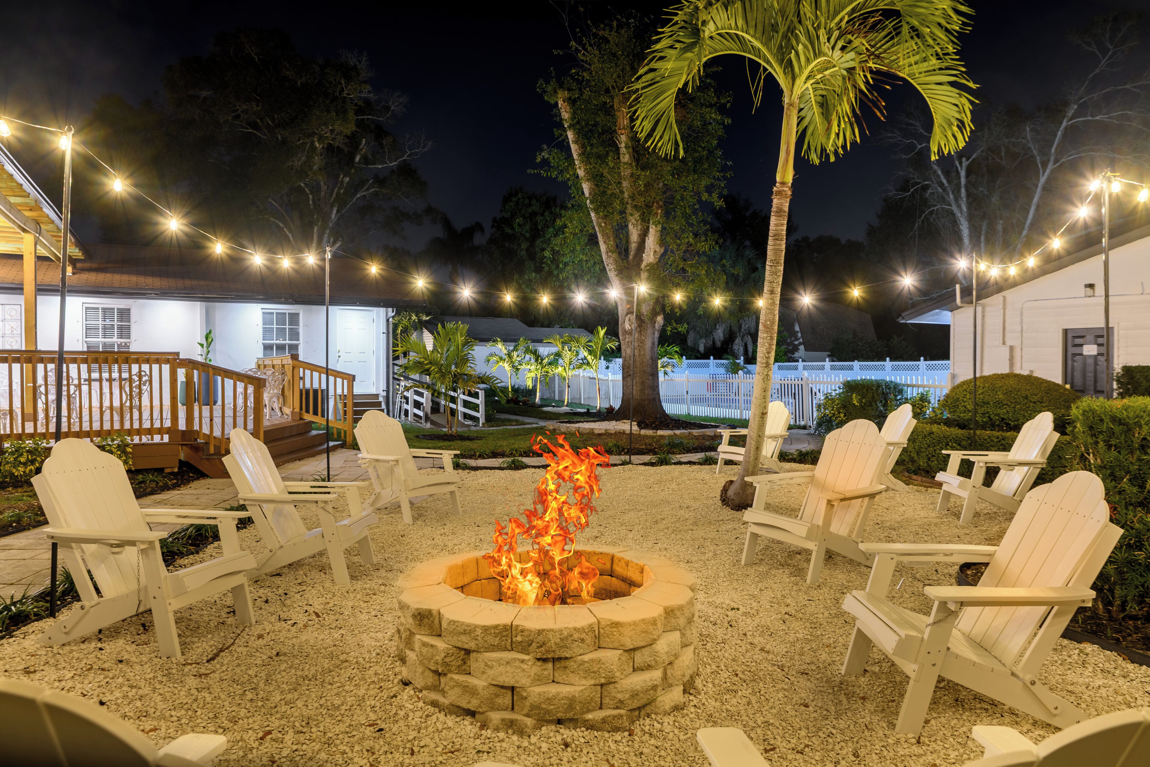 A fire pit surrounded by Adirondack chairs in a garden area with string lights, palm trees, and a building with a white exterior.