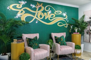 Quarto com duas poltronas cor-de-rosa, uma parede verde com a palavra 'Love' escrita em dourado e uma mesinha lateral amarela com livros e uma planta.