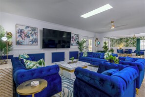 A modern living room with blue sofas, a flat-screen TV, and tropical-themed artwork. - 12 BR Estate!•HeatedPool • Pickle • GameRm •Events (Clearwater)