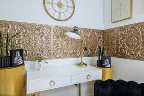Ett vitt skrivbord med guldfärgade detaljer, en svart soffa, dekorativa väggpaneler, en guldfärgad klocka och inramade konstverk.