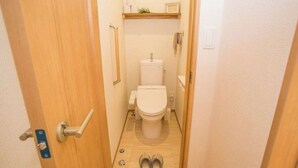 A narrow hallway leading to a toilet with a wooden door, a small shelf above the toilet, and a pair of slippers on the floor. - Resort House Daisho G Uruma (Uruma)