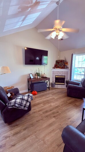 A living room with a fireplace, a ceiling fan, a television, and a large window. - River Pointe Getaway - Pigeon Forge Home + River Access, 3 Mins to the Parkway (Pigeon Forge)
