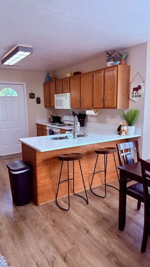 Una cocina con armarios de madera, encimera blanca, microondas y lavamanos. Hay dos taburetes y una silla.