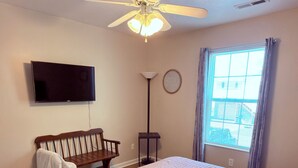 Un dormitorio con cama, ventilador de techo, televisor, banco, lámpara de pie y ventana.