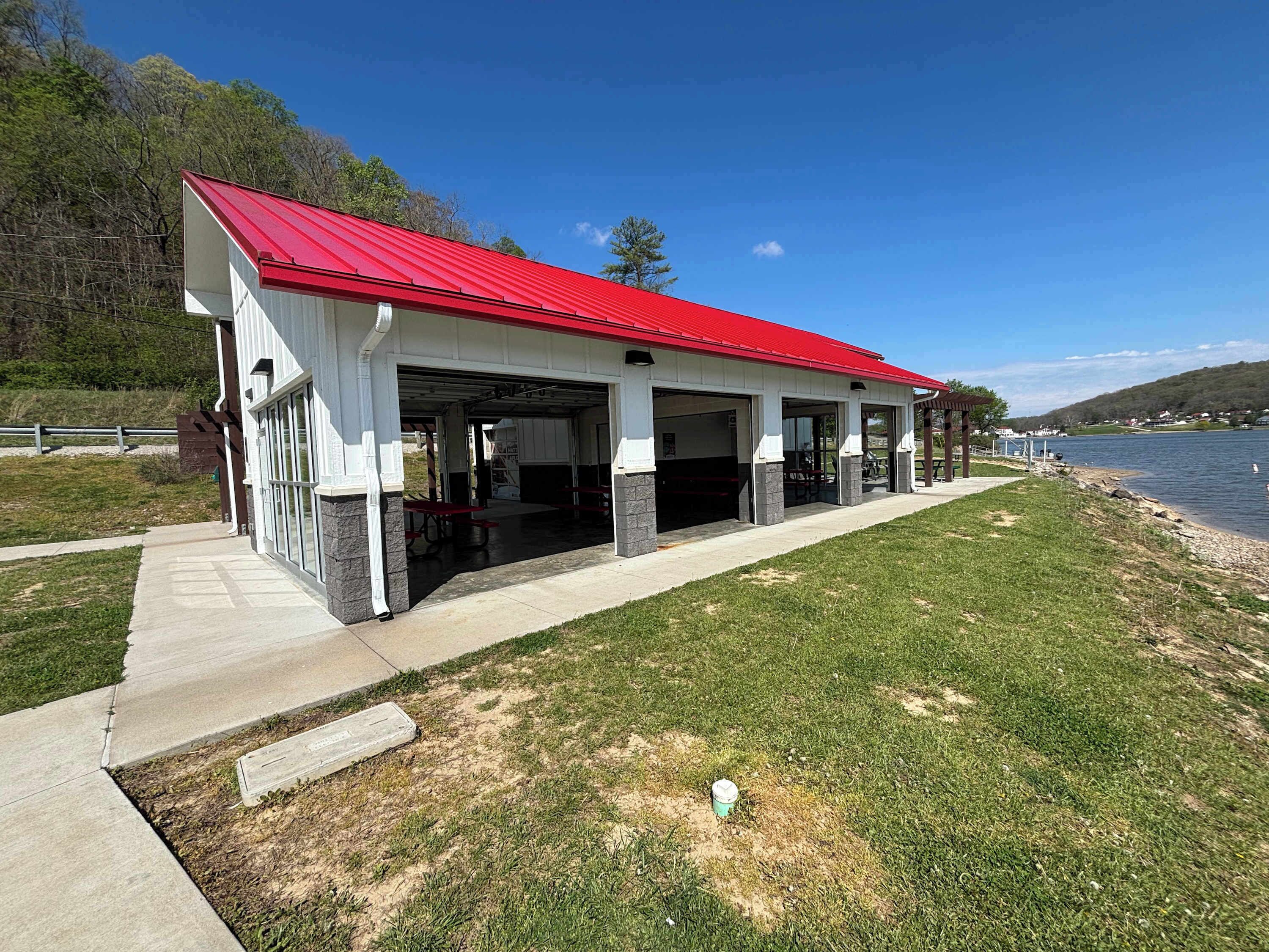 A covered outdoor area with a red roof, open garage doors, and a concrete pathway leading to a grassy area by a body of water.