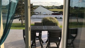 Blick aus einem Zimmer mit Tisch und Stühlen auf ein weißes Gebäude mit rotem Dach, umgeben von Grün und blauem Himmel.