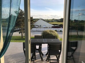 Blick aus einem Zimmer mit Tisch und Stühlen auf ein weißes Gebäude mit rotem Dach, umgeben von Grün und blauem Himmel.