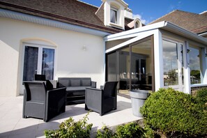 Een terras met buitenmeubilair, een glazen overkapping en een wit gebouw.