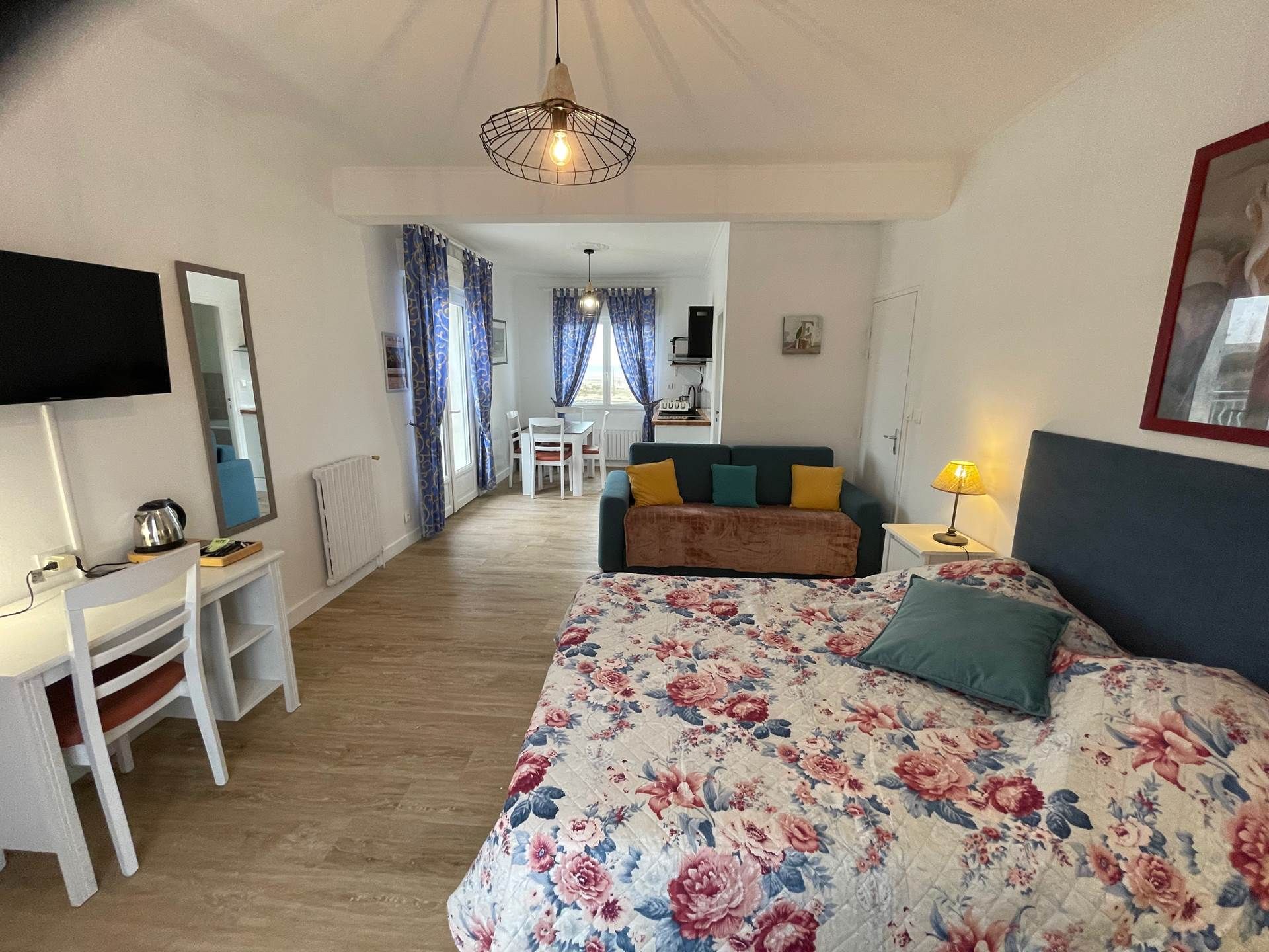 Un dormitorio con cama, escritorio, silla, televisión y una kitchenette al fondo.