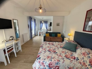 Ein Schlafzimmer mit Bett, Schreibtisch, Stuhl, Fernseher und einer kleinen Küchenzeile im Hintergrund.