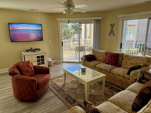 Ein Wohnzimmer mit Sofa, Sessel, Couchtisch, Fernseher und einem Balkon mit Gartenmöbeln.