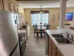 A kitchen with a refrigerator, stove, sink, dining table with chairs, and a window with blinds.
