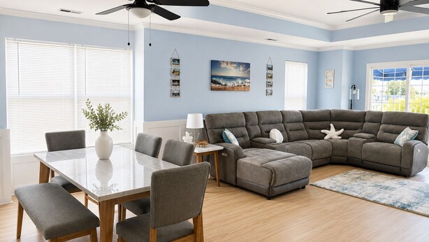 A living room with a sectional sofa, a dining table with chairs, a ceiling fan, and a large window. - New 3BR Wildwood Condo • Sleeps 8 • Garage • Walk to Beach (Wildwood)