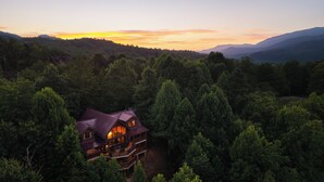 Un grand chalet à étages, doté d’un balcon imposant, entouré d’une forêt dense et de montagnes.