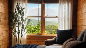 Une pièce de cabane en bois comprenant un lit, une chaise, une fenêtre donnant sur un paysage verdoyant et montagneux, et une plante.