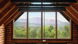 Une cabane en rondins dotée d’une grande fenêtre donnant sur un paysage montagneux boisé.